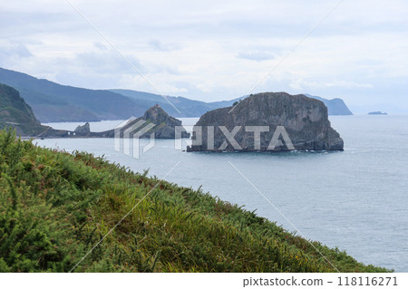 A cloudy daytime view of San Juan de Gaztelugatxe's church from Matxitxako Lighthouse, with rugged cliffs and greenery in the foreground. A cloudy daytime view of San Juan de Gaztelugatxe's church from Matxitxako Lighthouse, with rugged cliffs and greenery in the foreground. 118116271