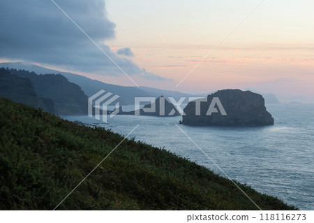 A serene dusk view of San Juan de Gaztelugatxe's church from Matxitxako Lighthouse, with rugged cliffs and lush greenery in the foreground. A serene dusk view of San Juan de Gaztelugatxe's church from Matxitxako Lighthouse, with rugged cliffs and lush greenery in the foreground. 118116273