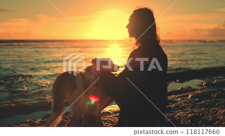 Woman sitting on beach with her dog enjoy bright colorful orange sunset over the sea. Girl female fondle her lovely pet. Feeling playful and carefree, relax, outdoor lifestyle summer holiday vacation Woman sitting on beach with her dog enjoy bright colorful orange sunset over the sea. Girl female fondle her lovely pet. Feeling playful and carefree, relax, outdoor lifestyle summer holiday vacation 118117660