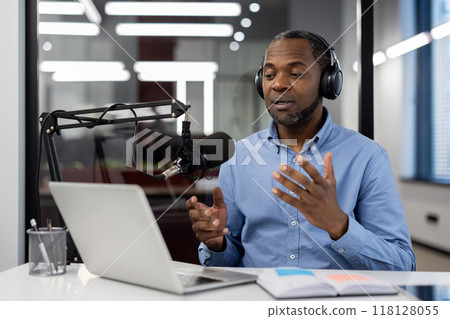 Man in office setting recording podcast, speaking into microphone with laptop. Wearing headphones for high-quality audio capture. Notebook and pen nearby for notes and ideas. Man in office setting recording podcast, speaking into microphone with laptop. Wearing headphones for high-quality audio capture. Notebook and pen nearby for notes and ideas. 118128055
