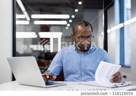 Business professional concentrating on reviewing documents at desk with laptop in modern office. Man appears focused and engaged, highlighting concept of productivity and attention to detail. 118128059