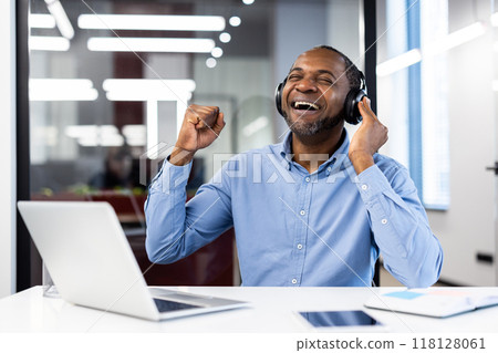 Smiling man in casual office setting wearing headphones, relishing music and celebrating achievement. Laptop and phone on desk highlight tech-driven work environment. 118128061