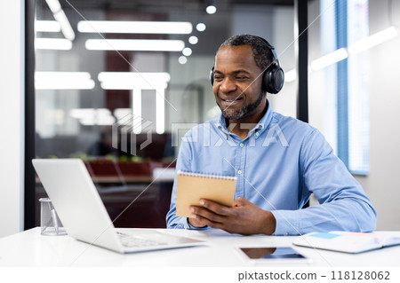 Businessman in blue shirt and headphones studies by watching webinar on laptop in office. He takes notes in notepad, demonstrating focus and engagement in professional development. Businessman in blue shirt and headphones studies by watching webinar on laptop in office. He takes notes in notepad, demonstrating focus and engagement in professional development. 118128062