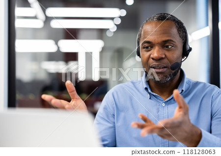 Confident professional man wearing headset in modern office engaged in video conference. Gesturing with hands, focused expression indicating thoughtful communication with team Confident professional man wearing headset in modern office engaged in video conference. Gesturing with hands, focused expression indicating thoughtful communication with team 118128063