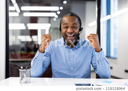 Joyful male office worker wearing headset celebrates success by raising fists. Smiling and filled with energy, he embodies achievement, motivation, and positive attitude in business setting. Joyful male office worker wearing headset celebrates success by raising fists. Smiling and filled with energy, he embodies achievement, motivation, and positive attitude in business setting. 118128064