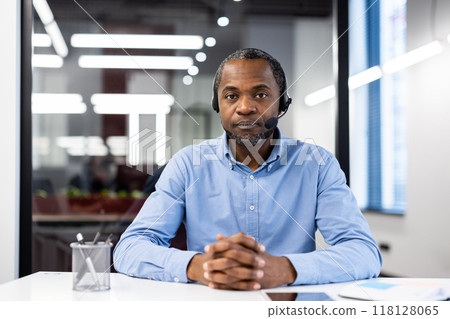 Professional businessman with headset at office desk communicates with clients. Confident expression conveys expertise. Office environment suggests corporate setting. Professional businessman with headset at office desk communicates with clients. Confident expression conveys expertise. Office environment suggests corporate setting. 118128065