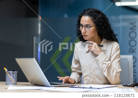 Hispanic businesswoman in formal shirt working on laptop, focused and serious in office setting. Concentrating on important tasks, holding pen, demonstrating professional mindset. Hispanic businesswoman in formal shirt working on laptop, focused and serious in office setting. Concentrating on important tasks, holding pen, demonstrating professional mindset. 118128671