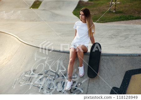 Attractive girl with a tattoo in a white t-shirt sitting on a ramp in skate park with a skateboard. 118128949