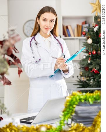 Doctor standing in medical office making notes on clipboard during christmas Doctor standing in medical office making notes on clipboard during christmas 118132303