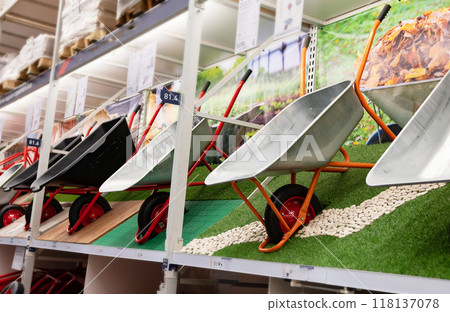 A young man with hands in gloves is carrying a metal garden cart through his beautiful green blooming garden. A professional gardener is carrying a wheelbarrow A young man with hands in gloves is carrying a metal garden cart through his beautiful green blooming garden. A professional gardener is carrying a wheelbarrow 118137078