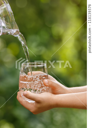 child hands holding glass and water pouring from jug with green garden on the background 118139376