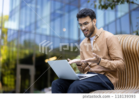 Man sitting outside on bench using laptop with confused expression, showing frustration and stress. Wearing casual clothes, gesturing with hands. Represents challenges of remote work Man sitting outside on bench using laptop with confused expression, showing frustration and stress. Wearing casual clothes, gesturing with hands. Represents challenges of remote work 118139393