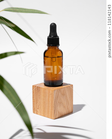 Face and body serum in bottle on a wooden cube podium on a white background with green leaves. Face and body serum in bottle on a wooden cube podium on a white background with green leaves. 118142143