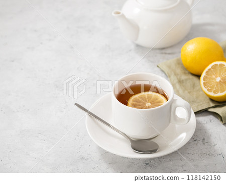 A cup of tea with lemon and spoon on a light background with a white teapot and citrus. A cup of tea with lemon and spoon on a light background with a white teapot and citrus. 118142150