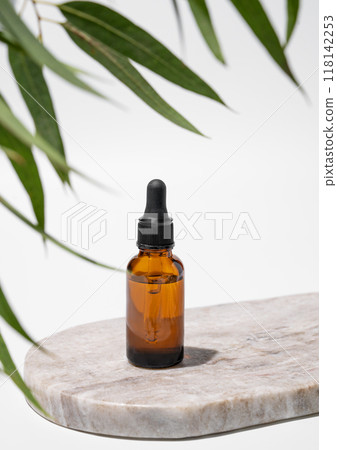 Face and body serum in bottle on a marble board on a white background with green leaves and shadow. 118142253
