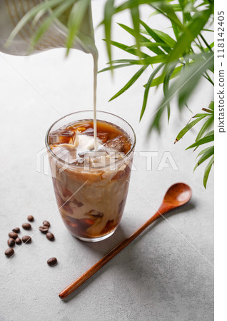 Iced coffee latte in a tall glass with a hand pouring milk from a milk jug on a light background  118142405
