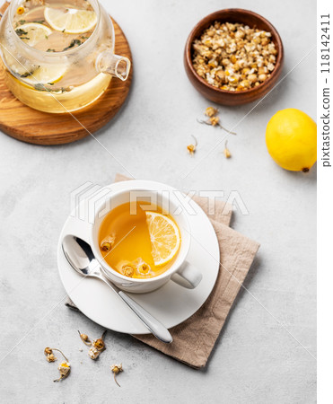 Chamomile herbal tea with lemon in a white cup and teapot with flowers on a light background. Chamomile herbal tea with lemon in a white cup and teapot with flowers on a light background. 118142411