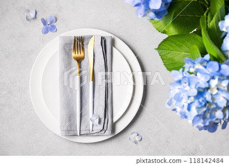 Festive table setting with blue hydrangea flowers and white plates on a light background. Festive table setting with blue hydrangea flowers and white plates on a light background. 118142484