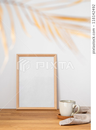 A frame with a blank canvas against a light wall and on a wooden tabletop with blurred palm leaf A frame with a blank canvas against a light wall and on a wooden tabletop with blurred palm leaf 118142492