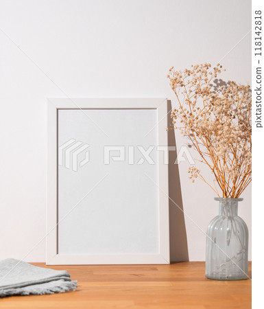 A white frame with a blank canvas against a light wall and on a wooden tabletop with vase A white frame with a blank canvas against a light wall and on a wooden tabletop with vase 118142818