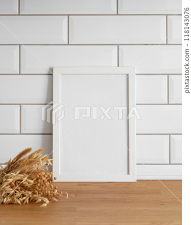 A  frame with a blank canvas against a white tile wall and on a wooden tabletop with dry bouquet. 118143076