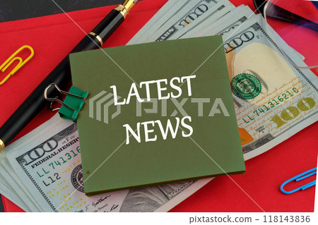 Conceptual latest news symbol. Copy space. LATEST NEWS message written on a green sticker on the background of dollar bills on the red book Conceptual latest news symbol. Copy space. LATEST NEWS message written on a green sticker on the background of dollar bills on the red book 118143836