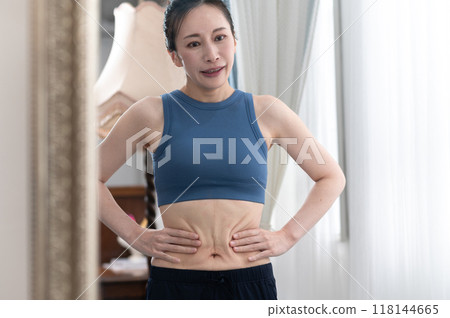 A woman in sportswear worries about her figure in front of a mirror A woman in sportswear worries about her figure in front of a mirror 118144665