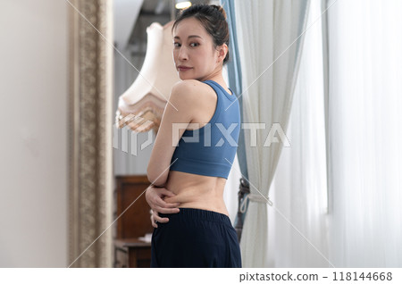 A woman in sportswear worries about her figure in front of a mirror A woman in sportswear worries about her figure in front of a mirror 118144668