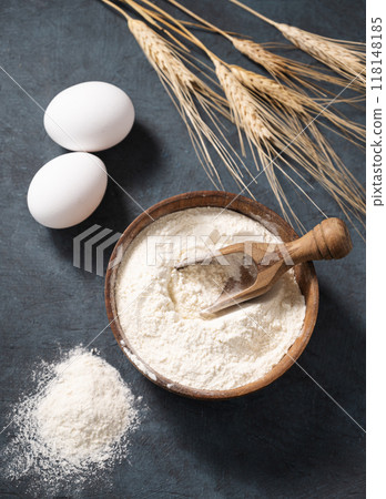 Wheat flour in a wooden bowl with a scoop on a dark blue background with eggs and dry branches wheat 118148185