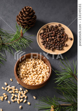 Pine nuts in a bowl and a scattered on a dark background with branches of pine needles and cone. 118149181