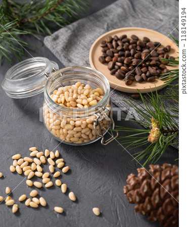 Pine nuts in a jar, bowl and a scattered on a dark background with branches of pine needles and cone Pine nuts in a jar, bowl and a scattered on a dark background with branches of pine needles and cone 118149194