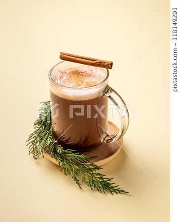 Glass mug of hot chocolate with whipped cream and cinnamon stick on a stand with pine branches Glass mug of hot chocolate with whipped cream and cinnamon stick on a stand with pine branches 118149201