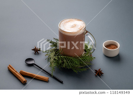Glass mug of hot chocolate with whipped cream and cinnamon stick on a stand with pine branches Glass mug of hot chocolate with whipped cream and cinnamon stick on a stand with pine branches 118149349