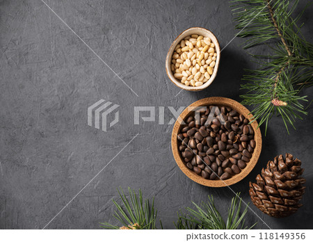 Pine nuts in a bowl and a handful of unpeeled nuts on a gray background with a branch Pine nuts in a bowl and a handful of unpeeled nuts on a gray background with a branch 118149356