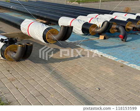 Stacked black pipes with curved ends, partially wrapped in insulation, on a paved construction site 118153599