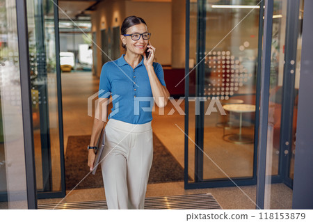A stylish businesswoman is exiting her office gracefully while on an important call A stylish businesswoman is exiting her office gracefully while on an important call 118153879