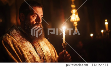Close up portrait of an Orthodox priest with a long beard wearing an ornate golden robe, holding a lit candle in a dimly lit church setting 118154511