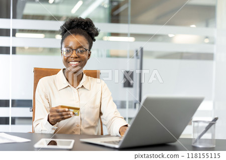 Confident African American businesswoman using laptop while holding credit card, symbolizing online shopping or e-commerce in professional office environment. Smartphone on table adds technological 118155115
