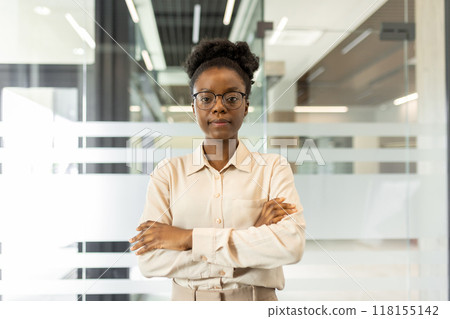 Confident African American businesswoman standing with arms crossed in modern office. Portrays leadership, confidence, and professionalism. Ideal for depicting successful business environment 118155142