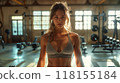 A young woman in a gym, standing confidently, preparing for a workout in a sunlit space 118155184