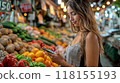 A woman shopping for fresh produce in a vibrant market, holding her phone while choosing vegetables 118155193