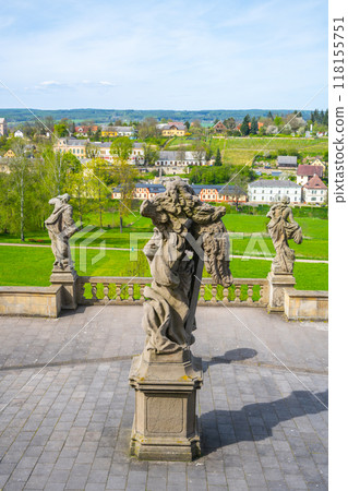 Baroque statues stand elegantly within the gardens of Kuks hospital complex, overlooking lush greenery and quaint buildings in the distance, creating a serene atmosphere. 118155751