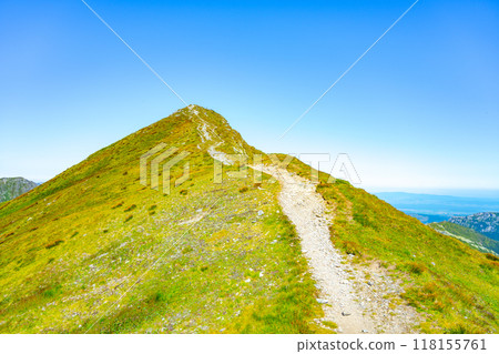 A winding hiking trail leads up Bystra Mountain in the Tatras, Slovakia. The landscape showcases vibrant green grass and a bright blue sky, inviting outdoor enthusiasts to explore. A winding hiking trail leads up Bystra Mountain in the Tatras, Slovakia. The landscape showcases vibrant green grass and a bright blue sky, inviting outdoor enthusiasts to explore. 118155761