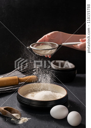 Wheat flour is sifted by a hand into a bowl on a dark background with eggs and a rolling pin. Wheat flour is sifted by a hand into a bowl on a dark background with eggs and a rolling pin. 118155788