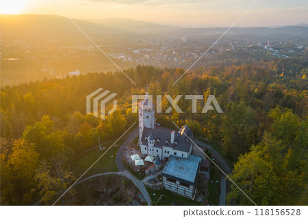 The Liberecka Vysina Lookout Tower stands majestically in Liberec, Czechia, surrounded by lush greenery as the golden sunset casts a warm glow over the landscape, inviting exploration and serenity. 118156528