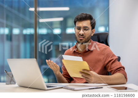 Young professional in office environment appearing frustrated after receiving bad news in postal envelope. Seated at desk with laptop, phone, and documents. Young professional in office environment appearing frustrated after receiving bad news in postal envelope. Seated at desk with laptop, phone, and documents. 118156600