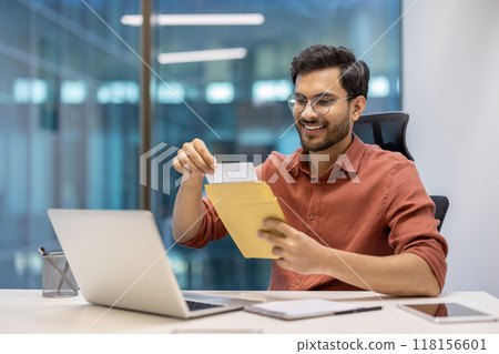 Young professional using laptop, opening envelope with joyful expression. Workplace setting with personal items, casual attire. Concept of work satisfaction, communication, modern technology. 118156601