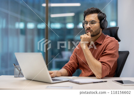 Serious businessman in modern office setting wearing headphones working on laptop. Focused on tasks, demonstrates concentration and professionalism. Represents technology use in business 118156602