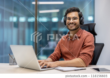 Young professional with glasses and headphones using laptop in modern office setting. Person smiling confidently while engaged in digital work. Office environment encourages creativity and focus 118156603