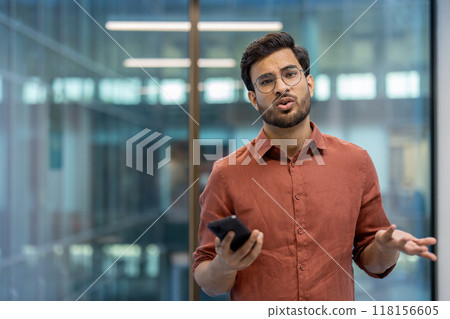 Dissatisfied upset man with phone in hands standing near office building, businessman looking at camera disappointed, read bad news online in social network. 118156605